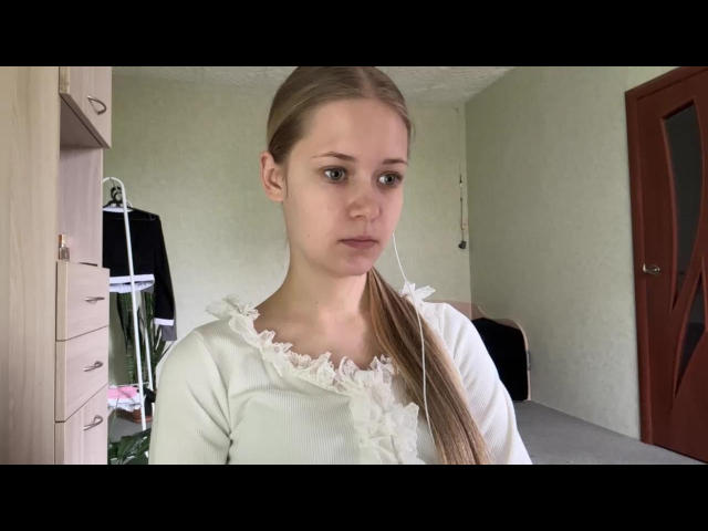 Lilliana Whang tem cabelo loiro preso em um rabo de cavalo, veste uma blusa branca com babados, aparenta seriedade, tem seios pequenos, pele clara e uma expressão neutra em um quarto modesto..