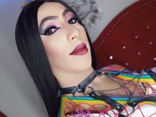 KattyDirty met zwart haar, roze oogschaduw, dikke wenkbrauwen, rode lipstick, draagt een top van regenboogkleurig gaas, een zwart harnas en kettingen, en laat haar borsten zien. Op de achtergrond is een rode hoofdbord te zien..