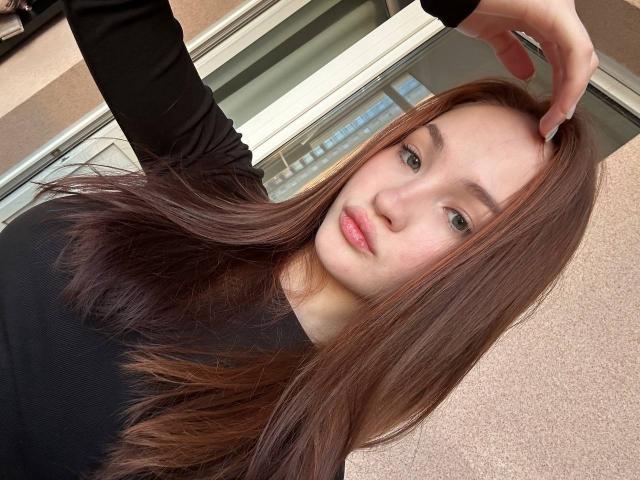 HanaGardinier ha lunghi capelli castani, pelle chiara e occhi verdi, indossa una maglietta nera, con la mano destra che tocca la testa, appare leggermente annoiata, in un selfie..