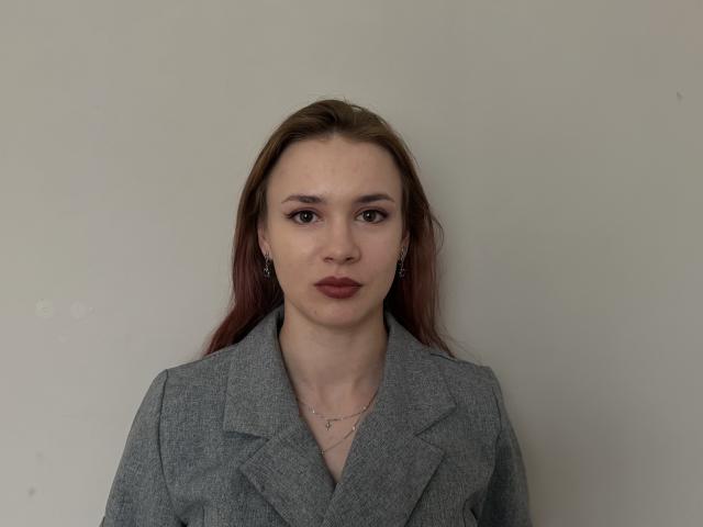 LeonaPapai with brown hair, wearing a gray blazer, has a neutral expression, red lipstick, and a small necklace, looking directly at the camera.