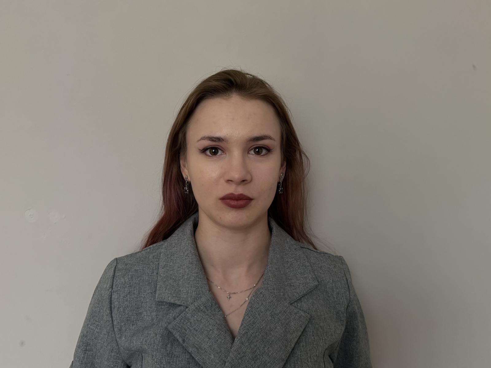 LeonaPapai, avec ses cheveux bruns, porte un blazer gris, a une expression neutre, du rouge à lèvres et un petit collier, et regarde directement l'appareil photo..