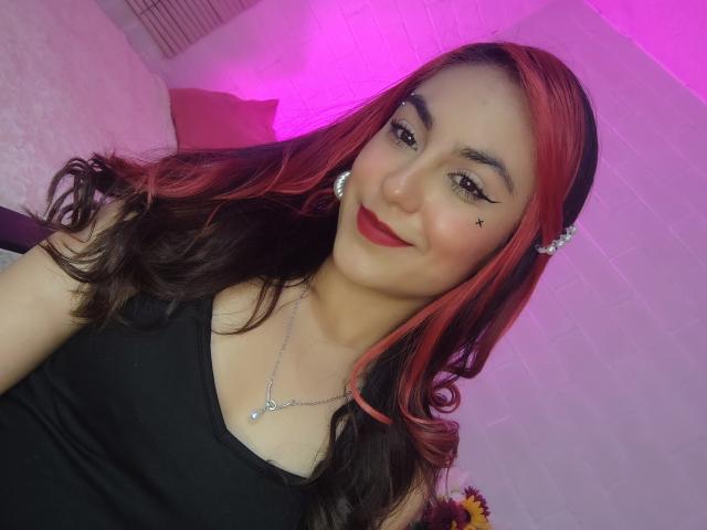 Amy, con cabello rojo y negro, delineador de ojos negro, lápiz labial rojo, camiseta negra, collar, sonriendo, iluminación rosa y morada, pared blanca y aretes. Se ven los senos debajo de la camiseta..