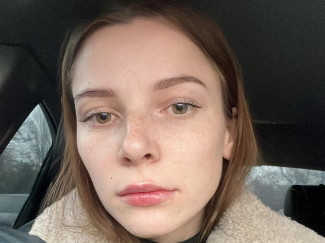 Il viso di Meliss69, con leggere lentiggini, occhi verdi e labbra rosa lucide, e capelli castani, indossa una giacca beige e soffice, ed è seduta in un'auto. Il suo naso è leggermente all'insù..