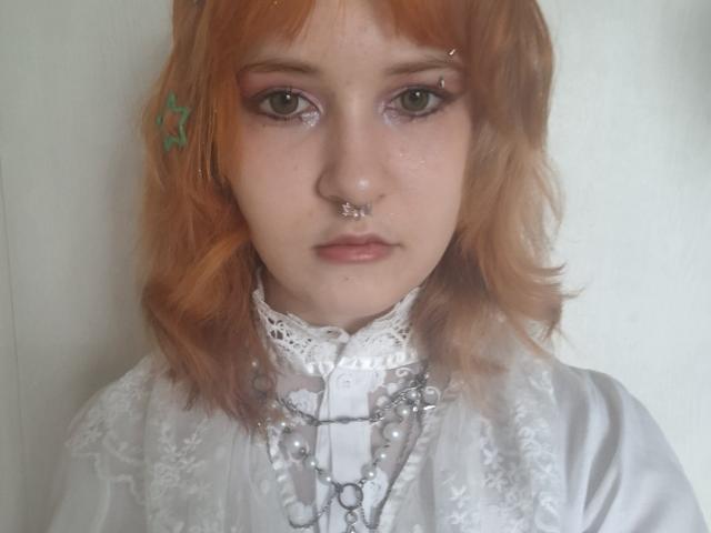 Taryn Mozzone mit orangefarbenem Haar, einem grünen Stern-Haarspange, einem Nasenring und einem Septumring, trägt ein weißes Schnürkleid, mehrere Halsketten und pinkes Augen-Make-up und wirkt ernst..