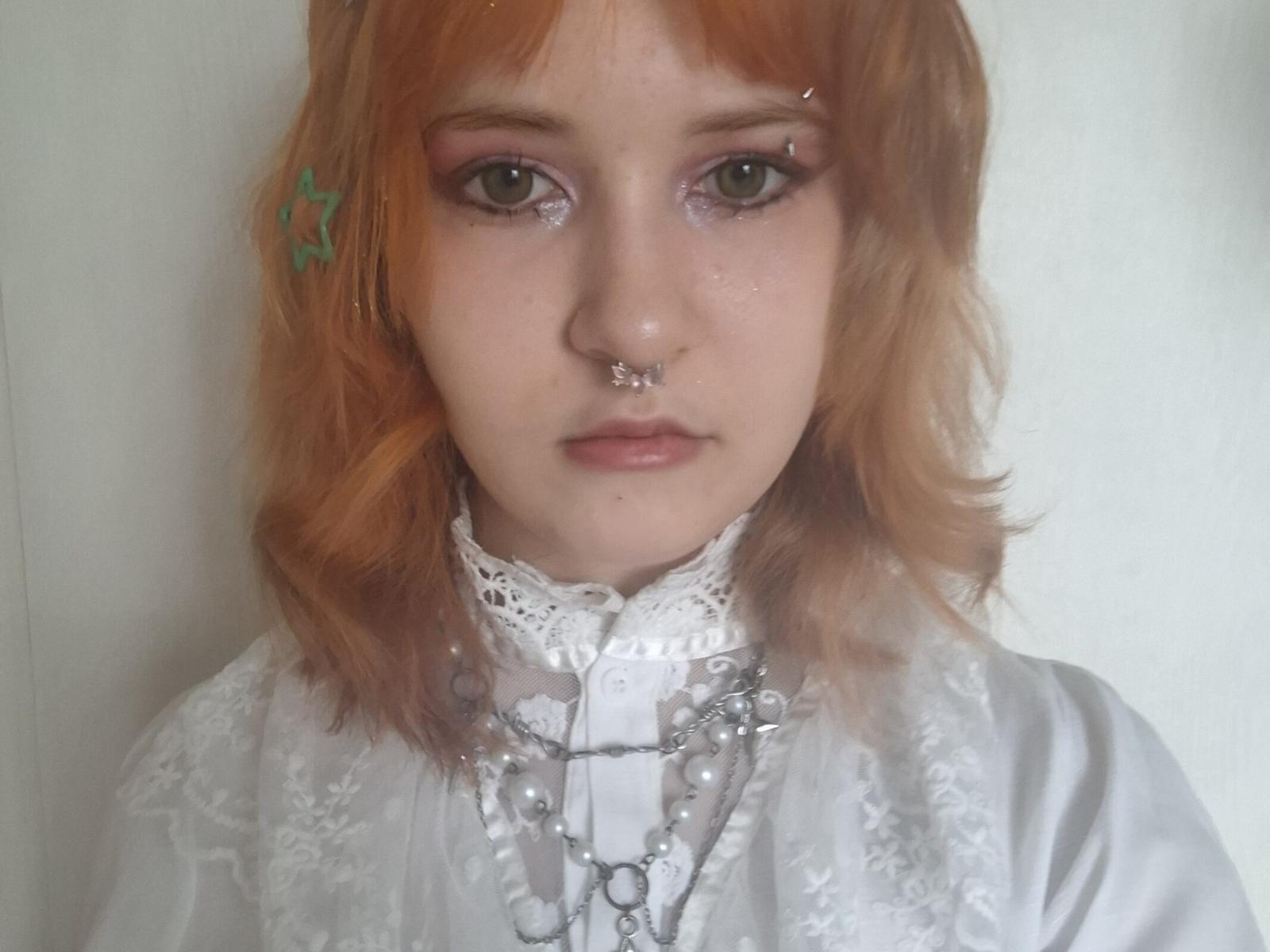 Taryn Mozzone, com cabelo laranja, um prendedor de estrela verde, um piercing no nariz e um piercing no septo, vestindo um vestido branco com cadarços, vários colares e maquiagem rosa nos olhos, com uma expressão séria..