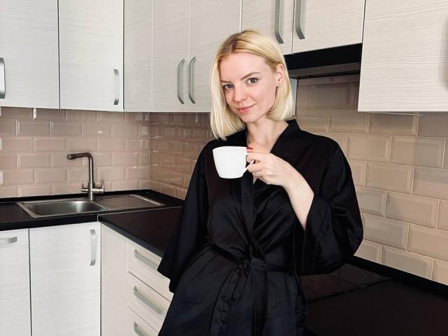 TeresitaBrugal with blonde hair, black silk robe, holding a white cup, standing in a modern kitchen, smiling subtly, hands in pockets, white tile backsplash, and white cabinets.
