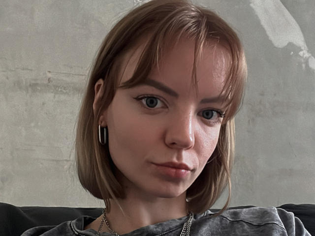 TeresitaBrugal has short brown hair, blue eyes, and a slight smile. She's wearing a gray shirt and small black earbuds with a nose ring. Her face is slightly flushed.