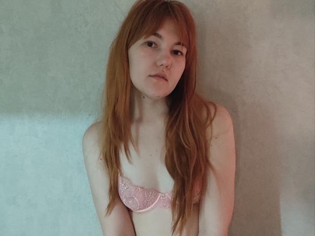 SheritaChieng with long red hair, pale skin, wearing a pink lace bra, looking down with a neutral expression, standing against a textured wall.