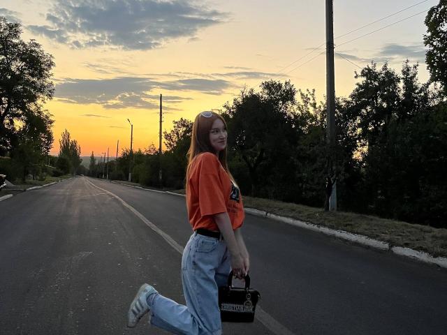 EstellVanderiet hat rotes Haar, ein orangefarbenes Hemd und blaue Jeans und steht auf einer Straße bei Sonnenuntergang, mit einer schwarzen Tasche und weißen Turnschuhen, und lächelt schief..