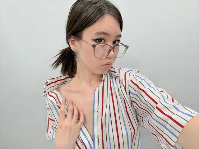 Emeline Lattig, con gli occhiali, una camicia a righe e piccoli seni, che tiene il colletto della camicia, con due trecce marroni, un piercing al naso, un'espressione seria, su sfondo bianco..