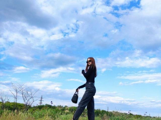 EstellVanderiet with long brown hair, wearing black top and pants, sunglasses, and black bag, standing in grassy field, blue sky with white clouds, looking to the side.