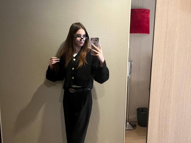 EstellVanderiet with long brown hair, black glasses, black cardigan, black high-waisted skirt, and black belt, taking a mirror selfie, holding phone in right hand, smiling.