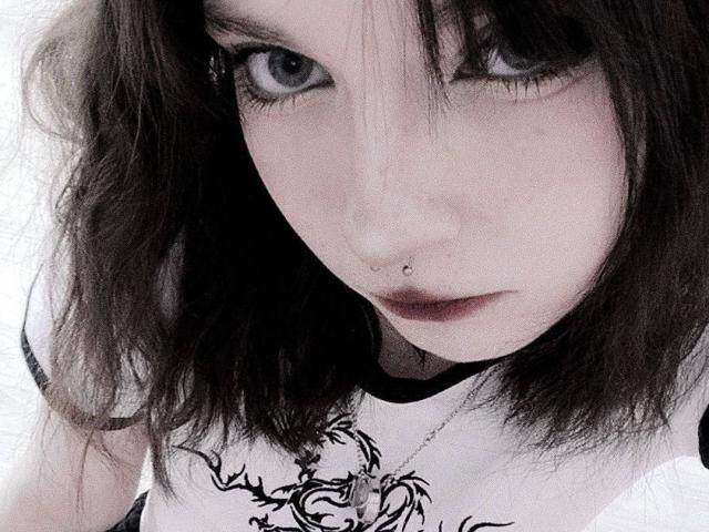 ElmaFasula's pale face, dark eyes, and messy brown hair with a nose piercing. She's wearing a black-and-white top with a decorative pattern. Her lips are slightly parted.