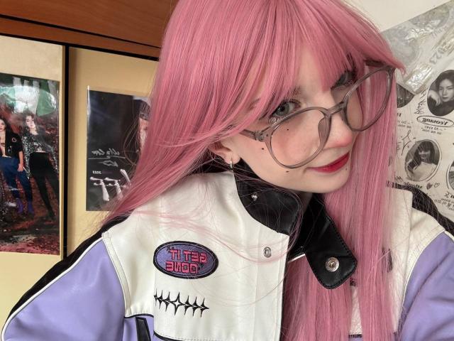 ElmaFasula with pink hair, glasses, and red lipstick, wearing a white and purple "Little Suu" jacket, smiling at the camera. Her tits are small, and she's cute.
