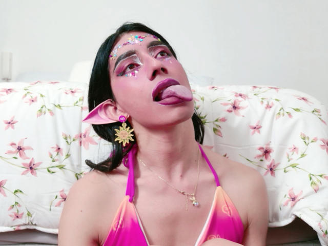 AlexaMissSx avec une peau rosée, des cheveux noirs, un fard à paupières rose, des paillettes, des oreilles pointues de type elfe, une langue rose en dehors, un bikini rose, des boucles d'oreilles dorées et un fond avec un lit de fleurs..