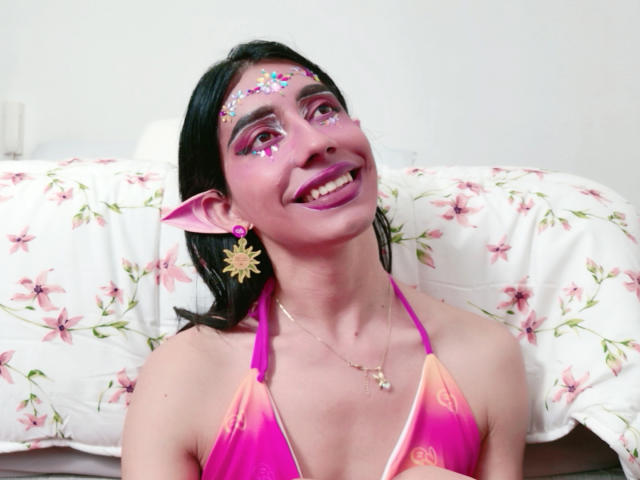 AlexaMissSx avec du maquillage rose, des paillettes sur le front, du rouge à lèvres violet, des oreilles pointues de lutin, un haut de bikini rose et orange, souriante, cheveux noirs, boucles d'oreilles en forme de soleil, lit avec une draps rose à motifs floraux..