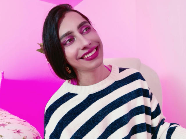 AlexaMissSx with short black hair, pink eyeshadow, and a smile, wearing a blue and white striped sweater, sitting in a pink-lit room, showing her teeth.