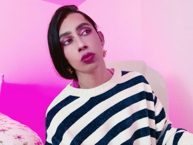 AlexaMissSx hat dunkles Haar, pinken Lidschatten, roten Lippenstift und trägt einen gestreiften, dunkelblauen und weißen Pullover. Sie sitzt in einem Raum mit pinker und violetter Beleuchtung. Ihre Brüste sind nicht sichtbar..