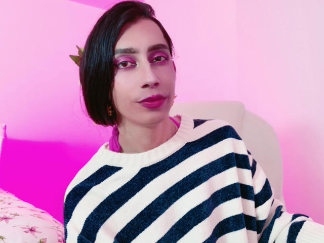 AlexaMissSx mit kurzen, schwarzen Haaren, rosa Lidschatten, lila Lippenstift, trägt einen weißen Pullover mit blauen Streifen und sitzt vor einem rosa beleuchteten Hintergrund, wobei sie leicht nach rechts schaut..