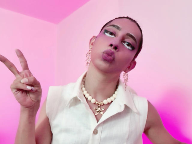 AlexaMissSx con trucco rosa, labbra carnose e medio levato, indossa una maglietta bianca senza maniche, una collana di perle e orecchini pendenti, sullo sfondo di pareti rosa..