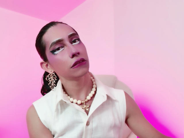 AlexaMissSx a les cheveux foncés attachés en queue de cheval, elle porte du maquillage blanc, un t-shirt blanc sans manches et un collier de perles. Elle regarde sur le côté, avec un éclairage rose et violet..