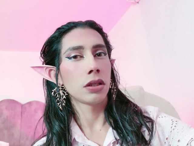 AlexaMissSx met puntige elfenoren, donker nat haar, roze eyeliner, gouden oorbellen en een lichtroze huid, gekleed in een wit shirt, met een licht verwarde blik..