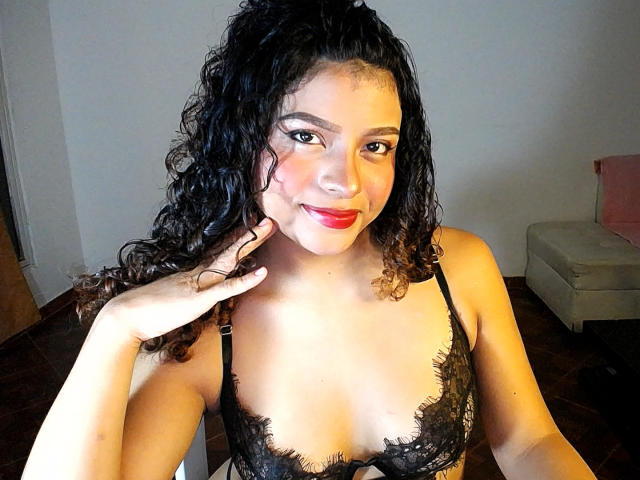 LuluPeach69X, avec des cheveux noirs bouclés, portant un soutien-gorge en dentelle noire, souriante, avec du rouge à lèvres rouge, une main sur la joue, seins de taille moyenne, peau brun clair, yeux foncés, assise dans une pièce faiblement éclairée..