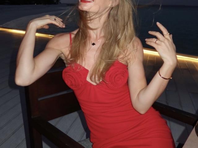 TheHottestBlonde ha lunghi capelli biondi, un vestito rosso con motivi floreali, rossetto rosso, sta sorridendo, ha le mani alzate, un anello al dito destro, un braccialetto al polso sinistro, ed è seduta su una sedia scura..
