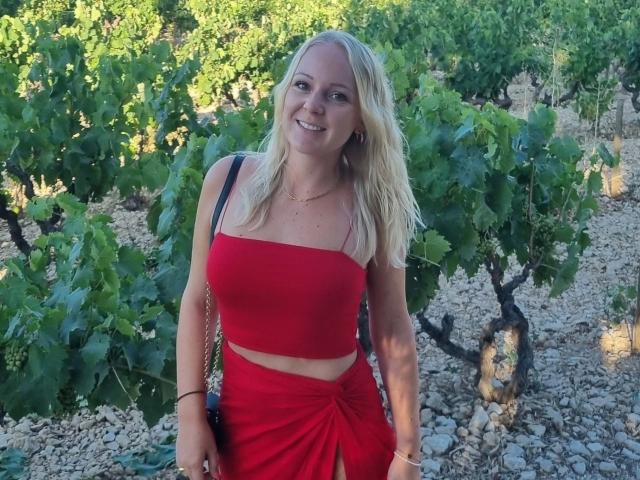 GermanMaskedLuci - Jolie femme blonde, souriante, portant un haut rouge à une épaule et une jupe assortie, laissant entrevoir un peu de ventre, avec un sac bandoulière noir, debout dans un vignoble..
