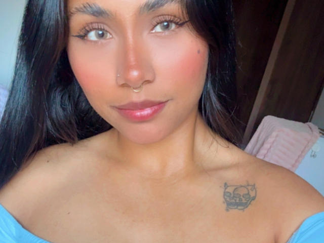 AnaRome with tan skin, long black hair, nose ring, subtle tattoo on right shoulder, blue off-shoulder top, pink lips, and brown eyes. Boobs not visible.
