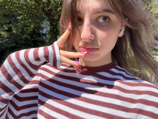 KarieMestler with striped shirt, brown eyes, pink lips, and long brown hair, touching face with pink nails, white earbuds, sunlight, and green trees in background.