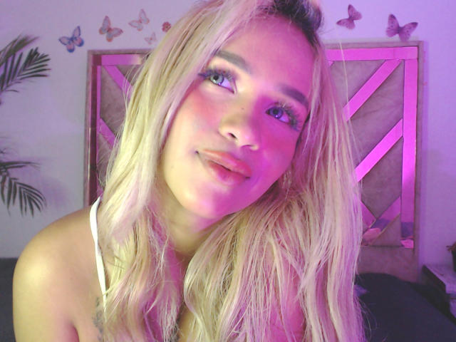 LuluDeVine mit blonden Haaren, blauen Augen und rosig-lila Beleuchtung, trägt ein weißes Oberteil, lächelt sanft, zeigt ein süßes Gesicht und eine kleine Nase, mit Schmetterlingen an der Wand..
