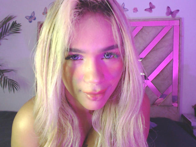 LuluDeVine with blonde hair, blue eyes, and a slight smile, showing her bare shoulders and a subtle lip piercing. Pink and purple lighting highlights her face.