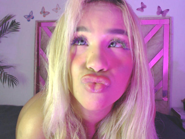 LuluDeVine has pink-tinted blonde hair, glossy pink lips, and green eyes with long, colorful eyelashes, pouting slightly, with purple and pink lighting on her face.