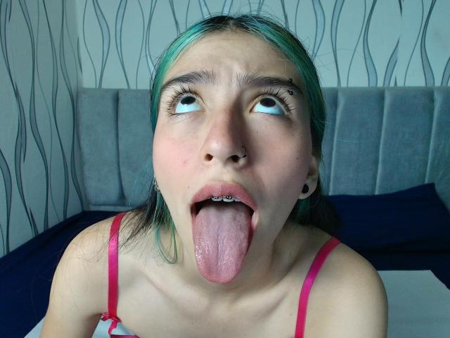 AbbyHillsI, met groen getint haar, blauwe ogen en een neusring, steekt haar tong uit, waardoor haar geperforeerde tong en zwarte oorbelgaten zichtbaar zijn. Ze draagt roze bandjes en kijkt omhoog..