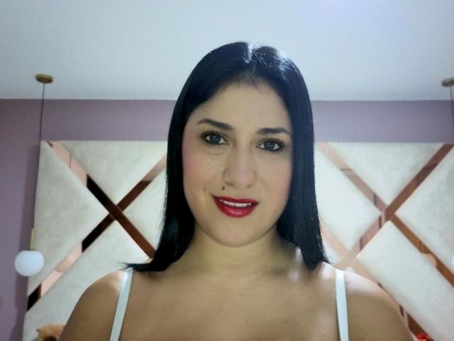 DamaraFoxy mit schwarzen Haaren, rotem Lippenstift, einem weißen Tanktop und sichtbaren Brüsten, lächelt in die Kamera und steht in einem Raum mit einem geometrischen, gepolsterten Kopfteil..