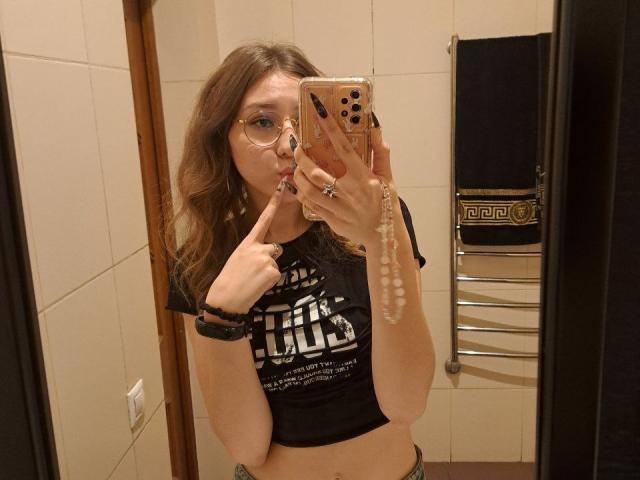 LenoreSchontz with wavy brown hair, glasses, black crop top, finger to lips, holding phone, pearl necklace, black bracelet, mirror selfie, tiled bathroom.
