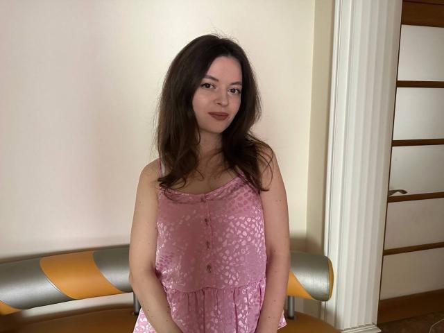 MartinaDuglas, con lunghi capelli castani, indossa un abito rosa, ricoperto di paillettes, senza maniche, ed è in piedi con le mani appoggiate sulla parte inferiore dell'addome, sorridendo leggermente, in una stanza con una panca gialla e grigia..