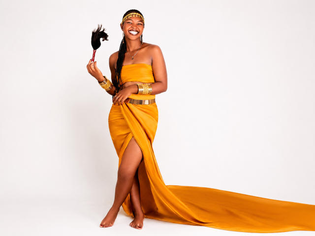 MelaninSugar stands barefoot, wearing a yellow strapless dress with a high slit, holding a black feathered fan, gold jewelry, and a headpiece, smiling with dark brown skin.