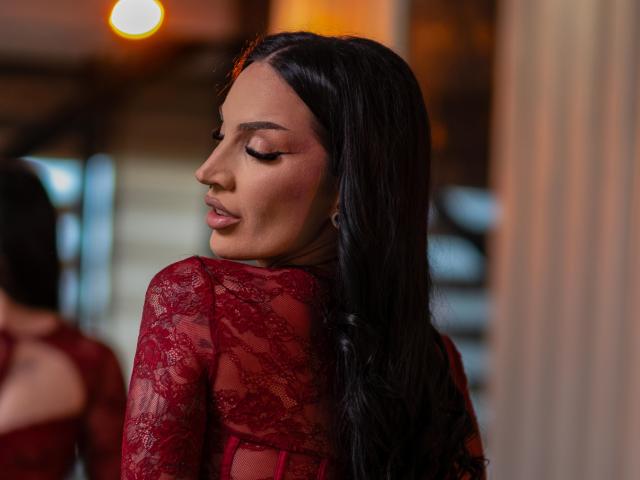 KyraReign, com seus longos cabelos pretos, olhos fechados e blusa de renda vermelha, exibe sua pele marrom lisa e maquiagem discreta. Seus lábios rosados e brilhantes estão ligeiramente entreabertos..