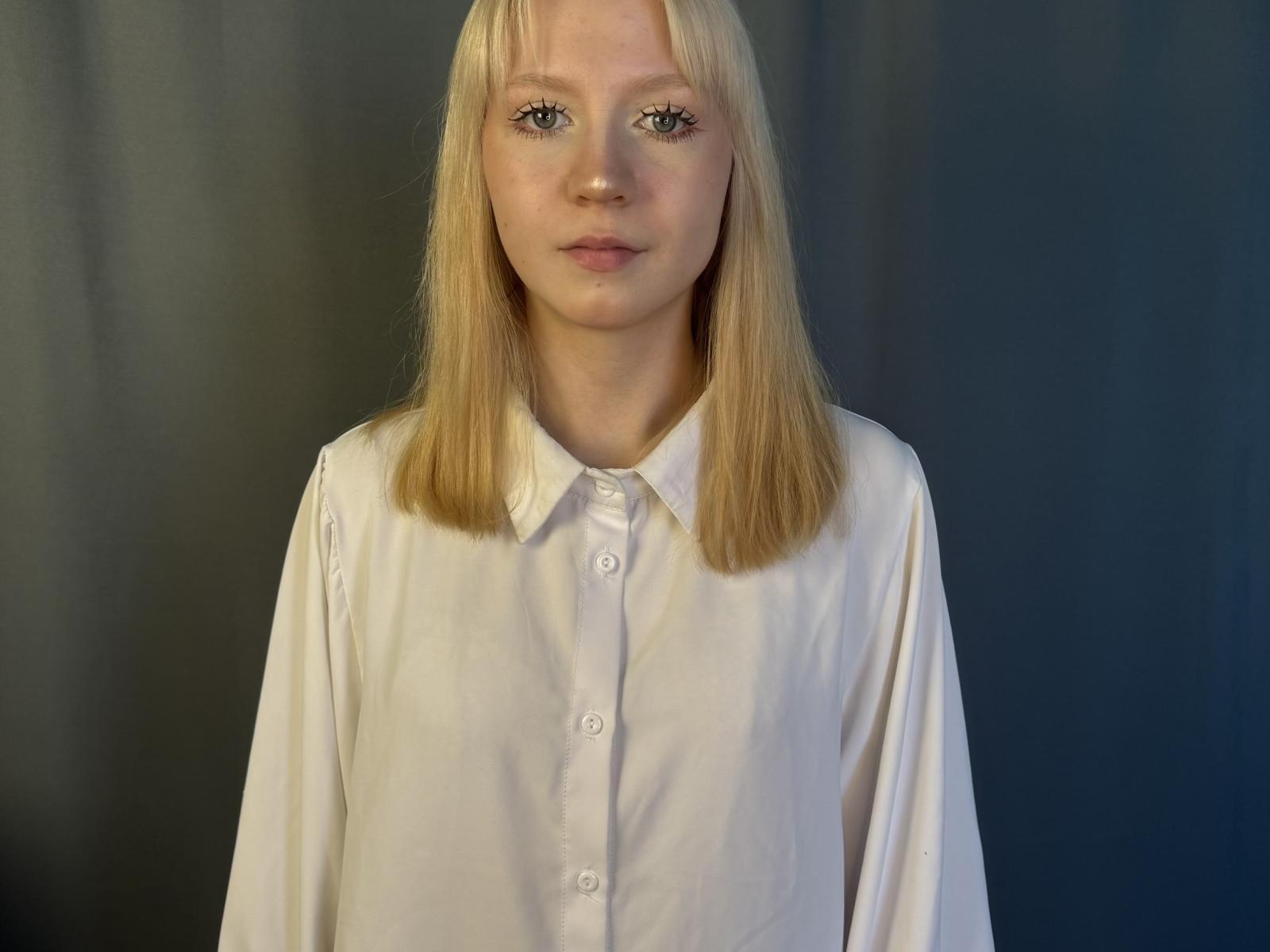 LaraineKnerien a les cheveux blonds et lisses, des yeux bleus, et porte une chemise blanche à boutons. Elle se tient devant un fond sombre et a une expression neutre..