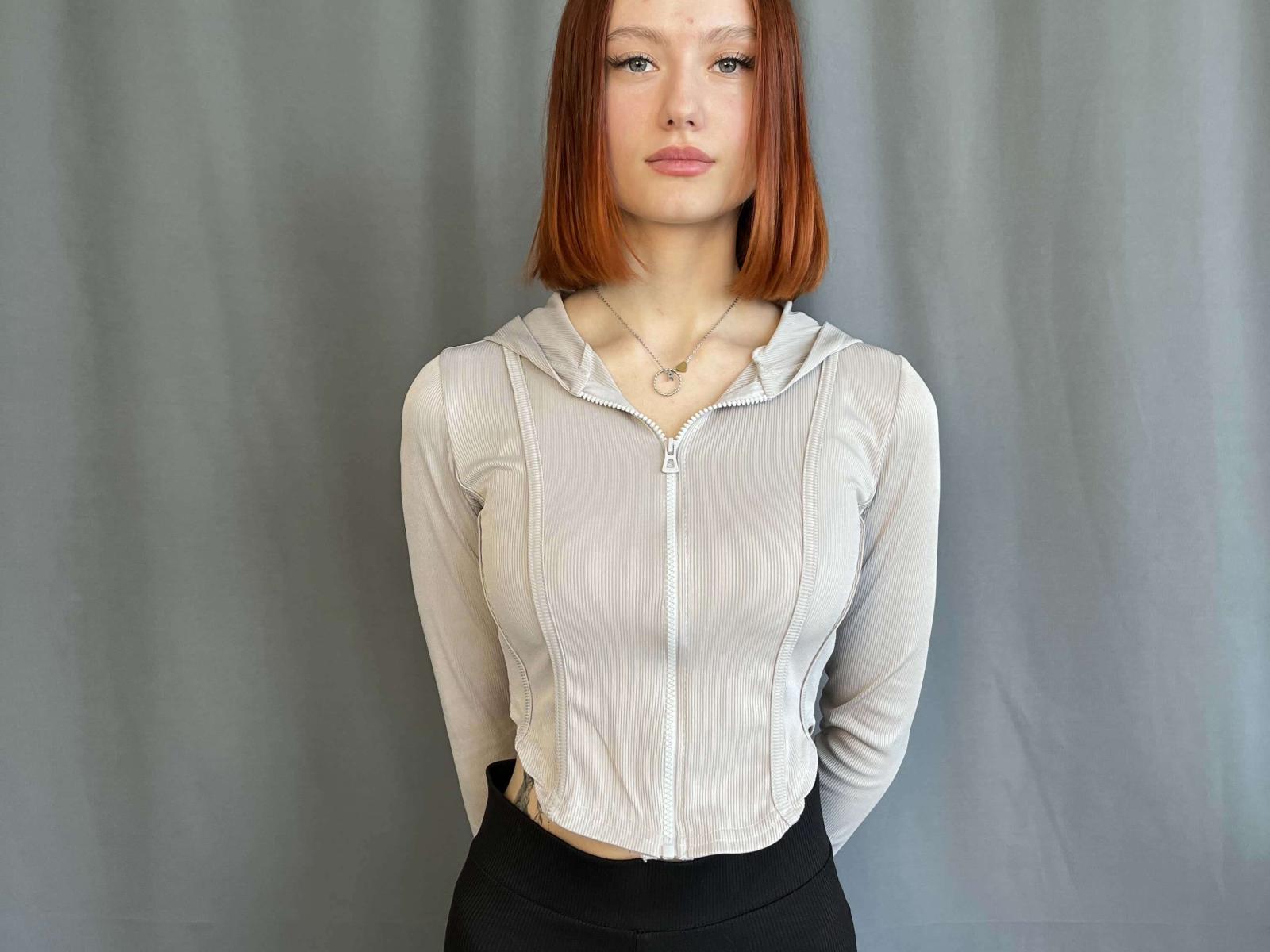 TaylorHersha with red bob, wearing a sheer white zip-up top showing her small tits, black skirt, and a necklace, standing against a gray background.