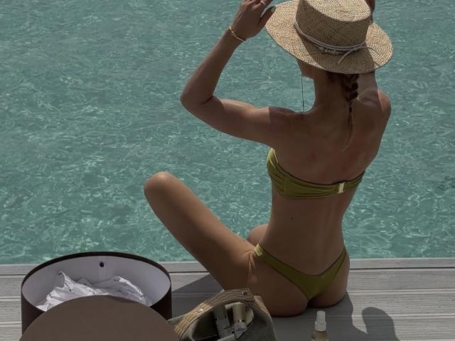 La rubia más atractiva, con un bikini verde, sentada de espaldas a la cámara, ajustándose un sombrero de paja. Su trasero bronceado y tonificado es visible. Un cubo y un sombrero están cerca..