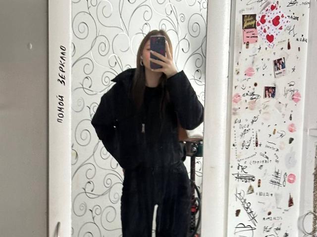 TericaSarley indossa una giacca nera, soffice e oversize, e sta scattando un selfie allo specchio con la mano in tasca, il telefono che le copre il viso, capelli biondi, sfondo bianco con motivi vorticosi..