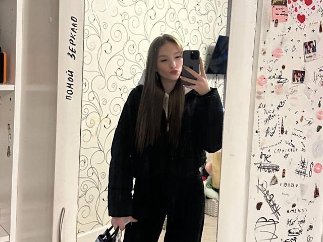 TericaSarley met lang bruin haar, een zwarte outfit, die een telefoon vasthoudt, een spiegelzelfportret, staand, met een lichte frons, die een zwarte tas vasthoudt, een witte muur met spiralen, rechts, een versierde muur aan de linkerkant..