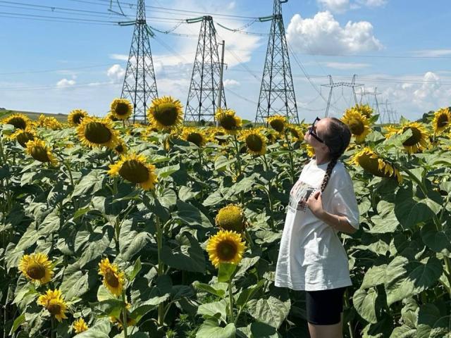 TericaSarley stands in a sunflower field, wearing a white shirt and black shorts, with a braid and sunglasses, looking up at tall electricity pylons on a bright day.