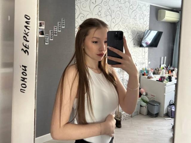 TericaSarley, avec de longs cheveux bruns, portant un haut blanc sans manches, tenant un téléphone pour un selfie devant un miroir, montrant ses petites seins et un léger ventre..