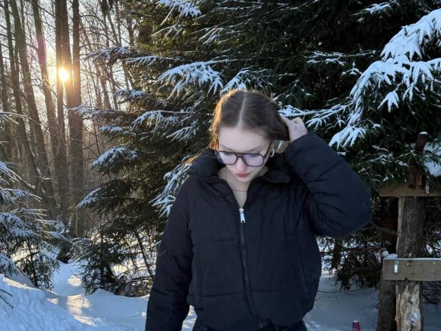 TericaSarley con gli occhiali, un giubbotto imbottito nero e le labbra rosse, in piedi nella neve, con una mano tra i capelli castani, il sole dietro gli alberi, abeti coperti di neve, foresta invernale, freddo..