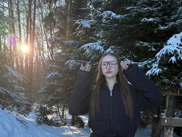 TericaSarley with long brown hair, glasses, and black puffy jacket, standing in a snowy forest, hands in jacket pockets, sun shining through trees, snow on pine branches.