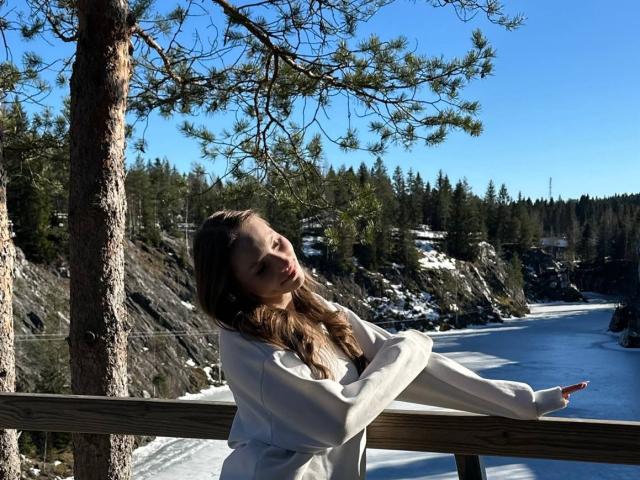 TericaSarley, con cabello castaño largo, ojos cerrados, apoyada en una barandilla de madera, vistiendo una sudadera blanca, de pie frente a un paisaje nevado, bordeado de pinos, con aguas azules, en un día soleado..